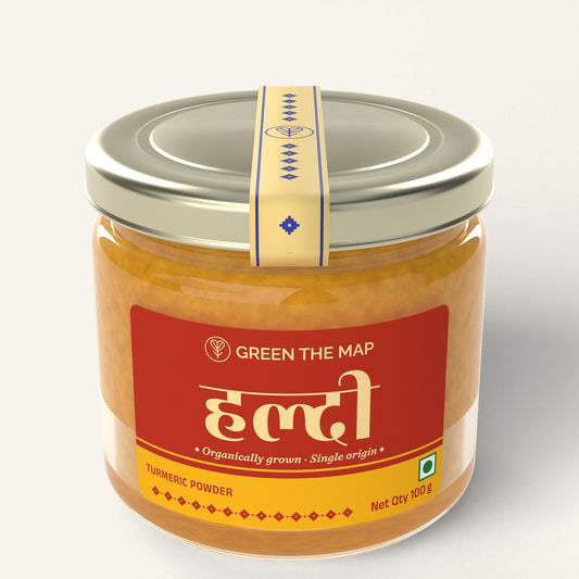 Turmeric powder 100 g in a glass jar with a red label and a champagne gold cap, on a plain off-white background.